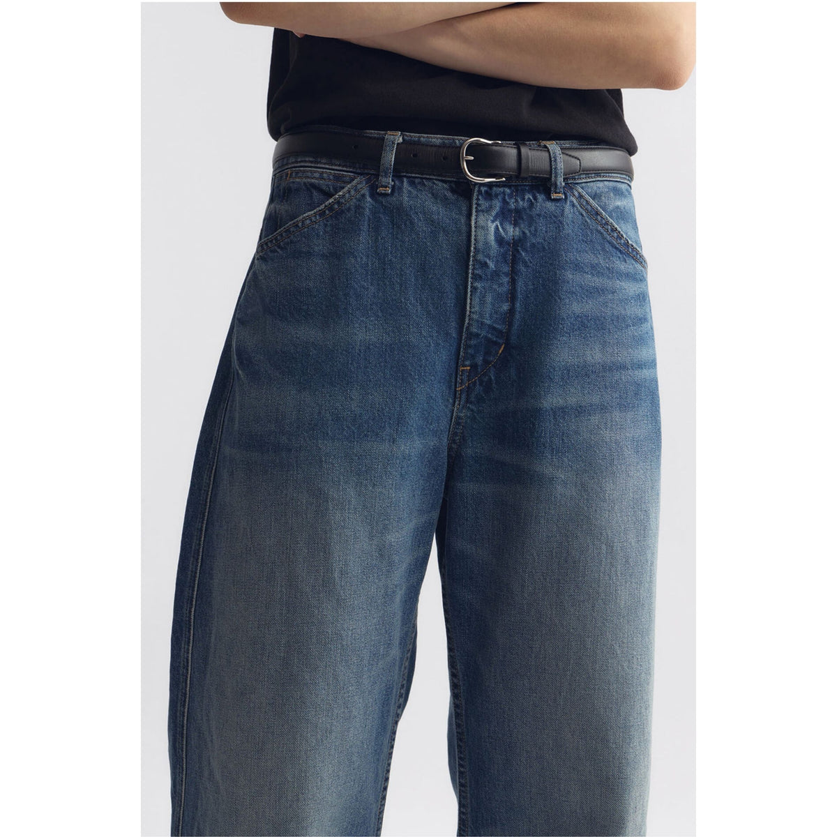 A close-up of a person wearing blue denim jeans with a black belt and black top, cropped from the chest down against a light gray studio background.