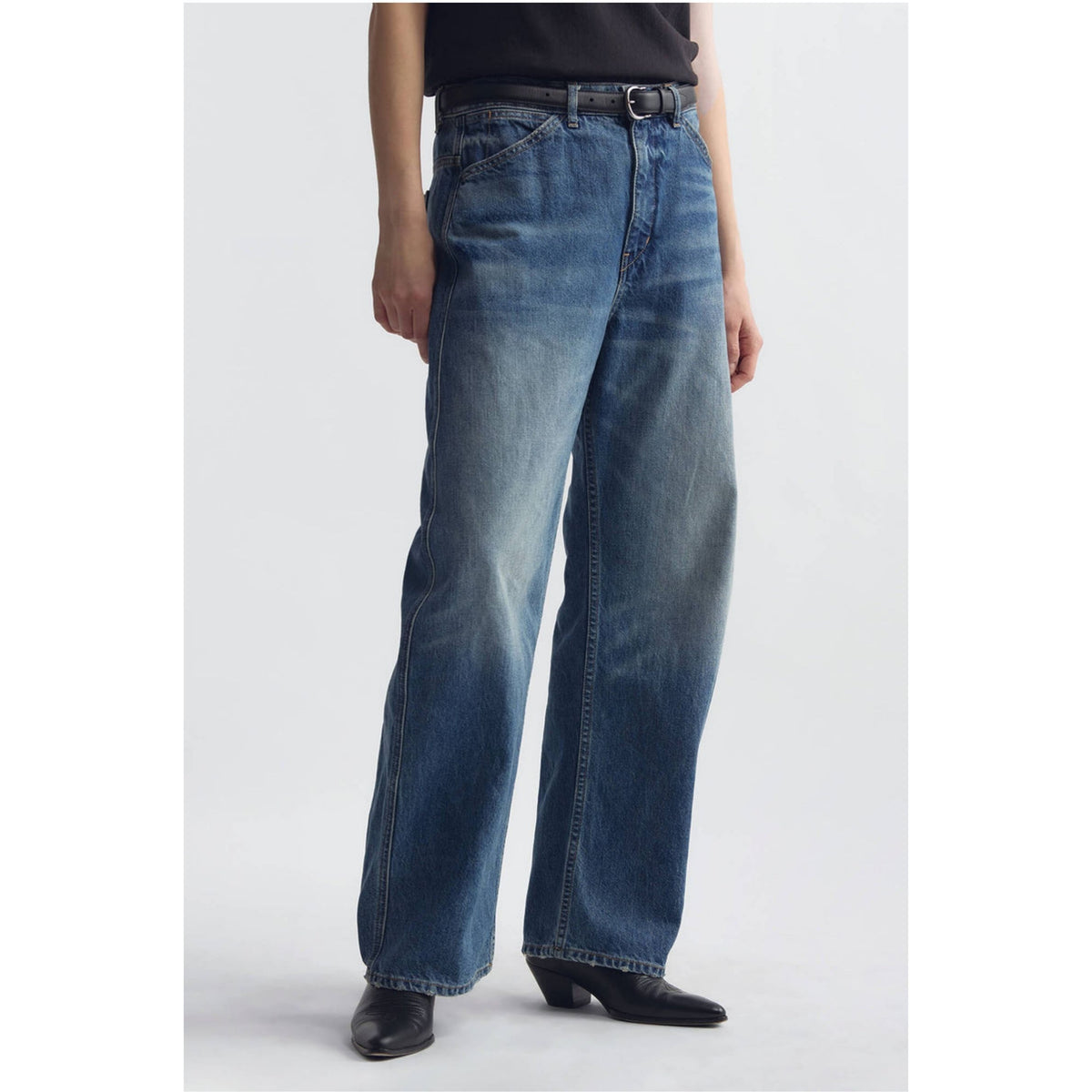 A model wearing a pair of wide-leg blue denim jeans with a faded wash, styled with a black belt, black top, and black pointed-toe heels against a light gray studio background.