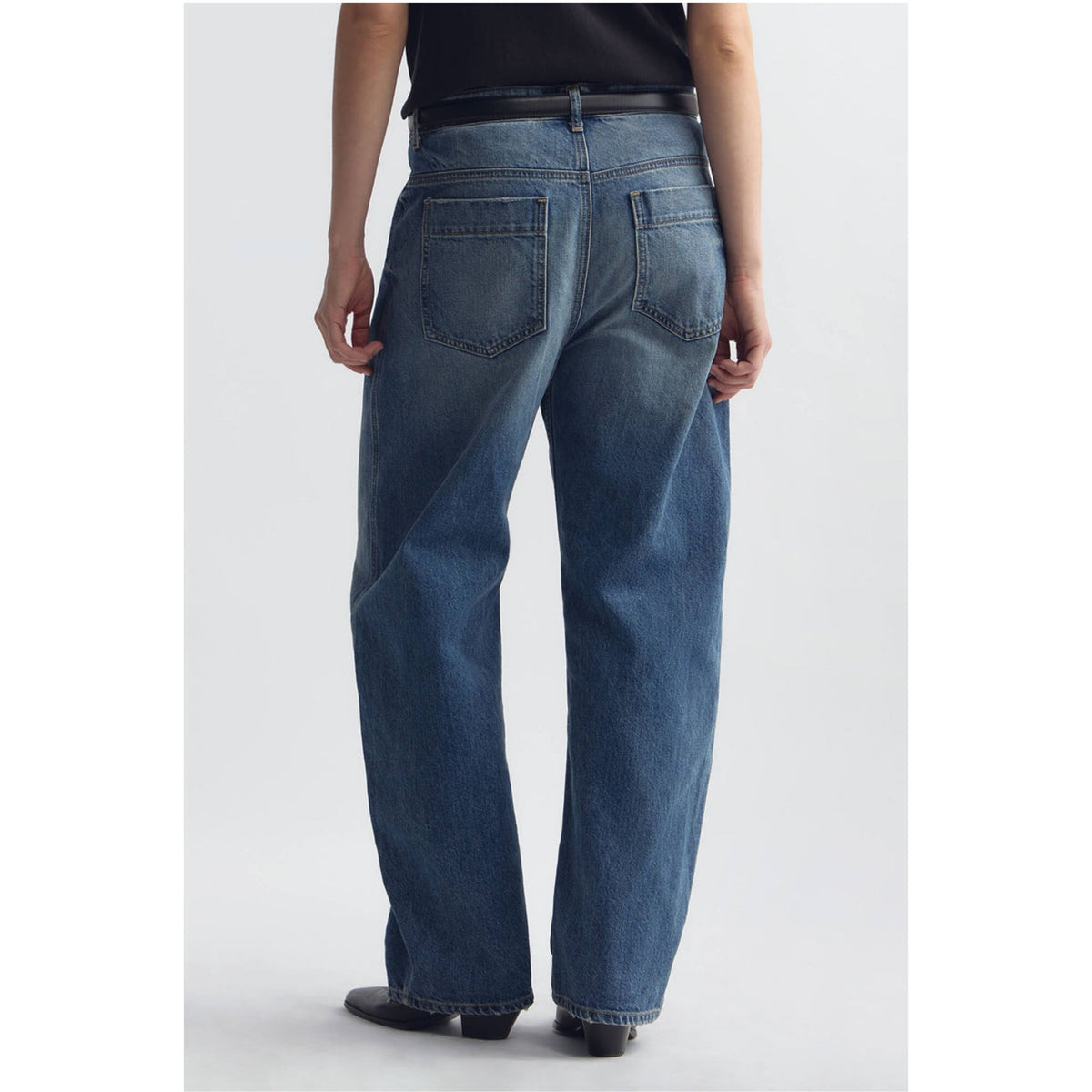 Rear view of a person wearing blue denim jeans with a black top and black shoes, standing against a light gray studio background.