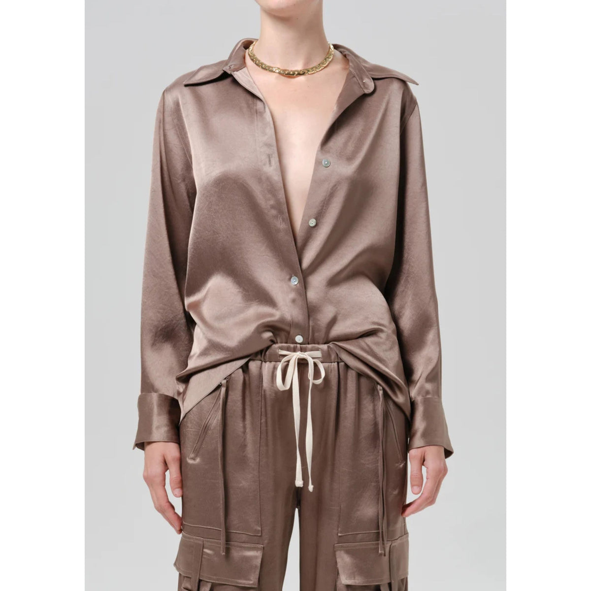 A model wearing a matching two-piece set consisting of a long-sleeve button-up shirt and drawstring cargo pants in a shiny taupe-brown satin-like fabric, styled with a gold chain necklace against a light gray background.
