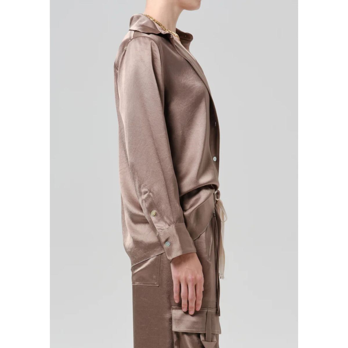 A side-profile view of a model wearing a shiny taupe-brown satin long-sleeve shirt and matching cargo pants against a light gray background.