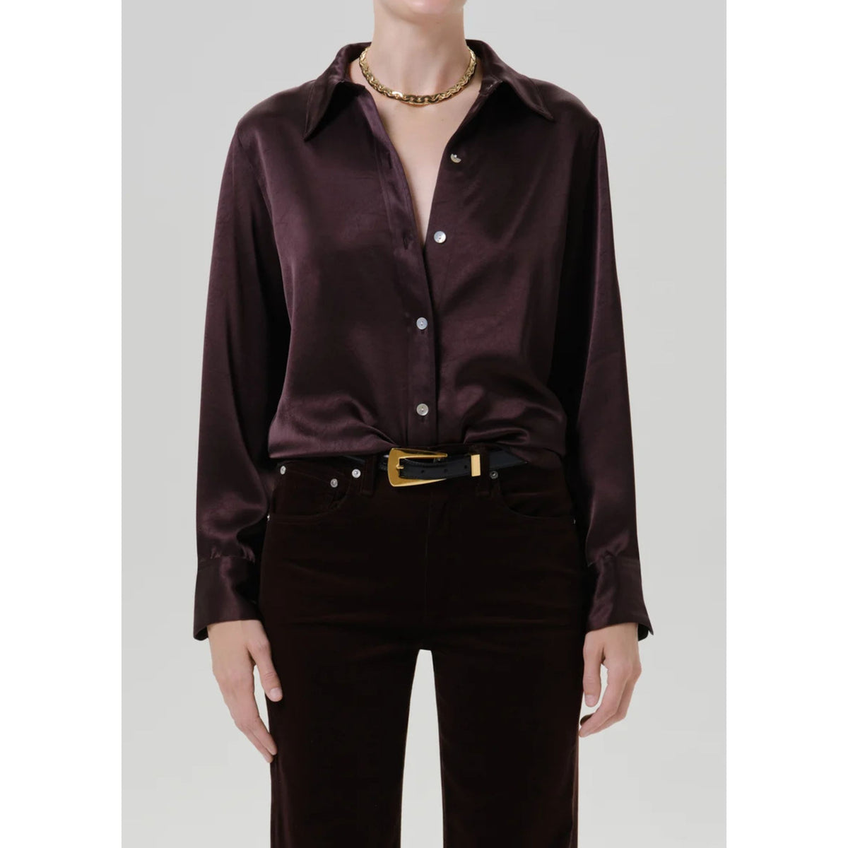 A model wearing a dark plum satin button-down shirt tucked into dark brown corduroy trousers, styled with a black belt with a gold buckle and a gold chain necklace, against a light gray background.