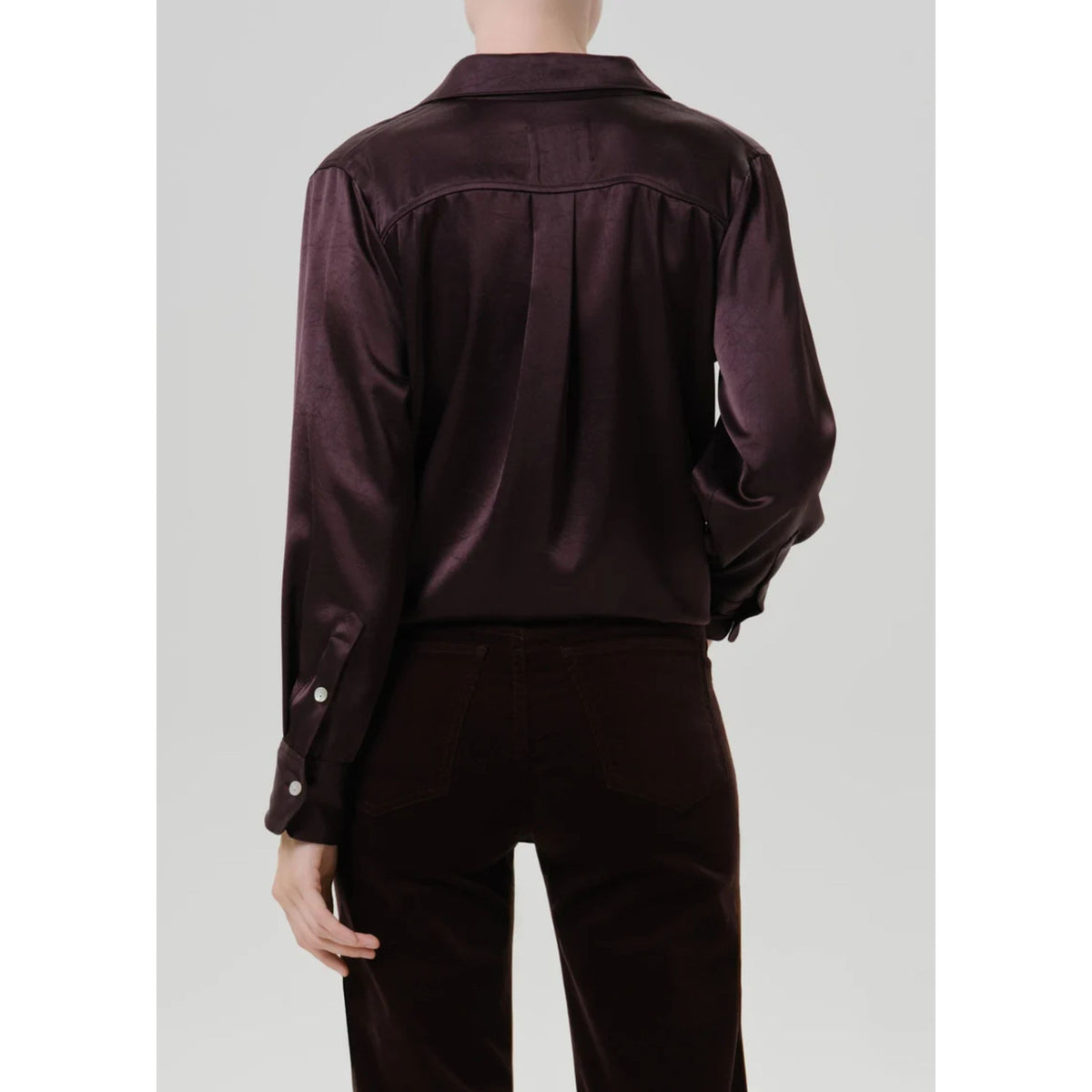 A model shown from the back wearing a dark plum satin shirt and matching dark brown corduroy trousers against a light gray studio background.
