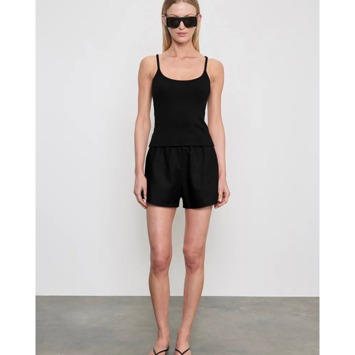 A woman modeling a black sleeveless camisole top and matching black shorts against a light gray studio wall and concrete floor.