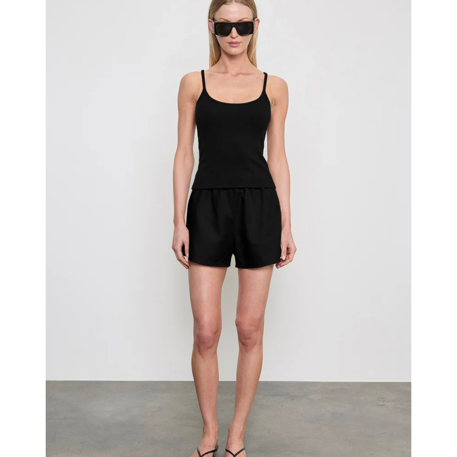 A woman modeling a black sleeveless camisole top and matching black shorts against a light gray studio wall and concrete floor.