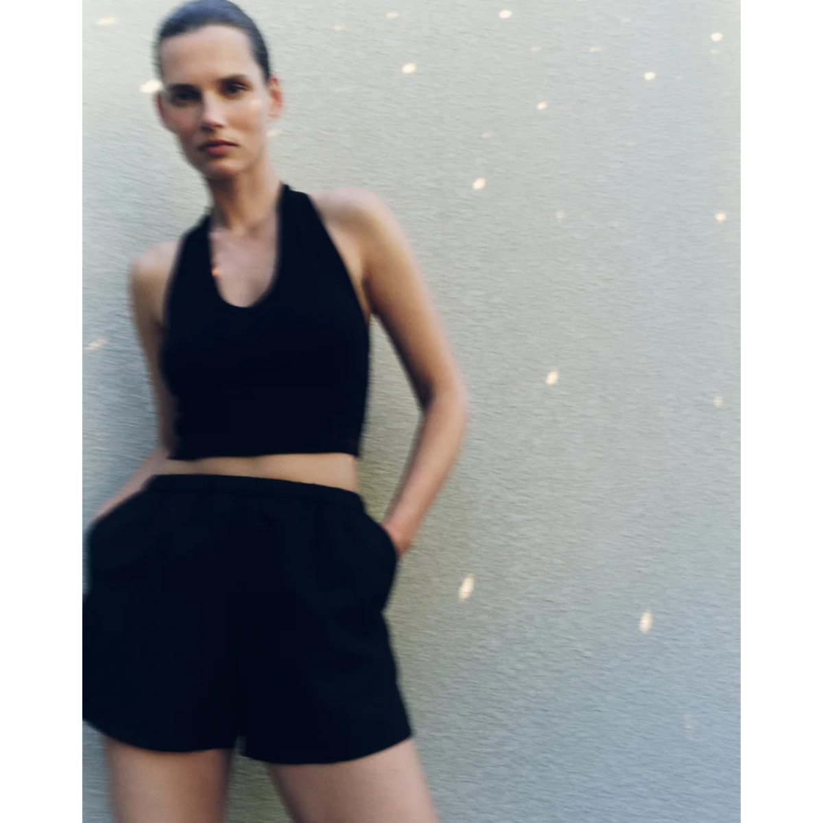 A woman wearing a black halter crop top and black shorts, standing against a textured light gray wall.