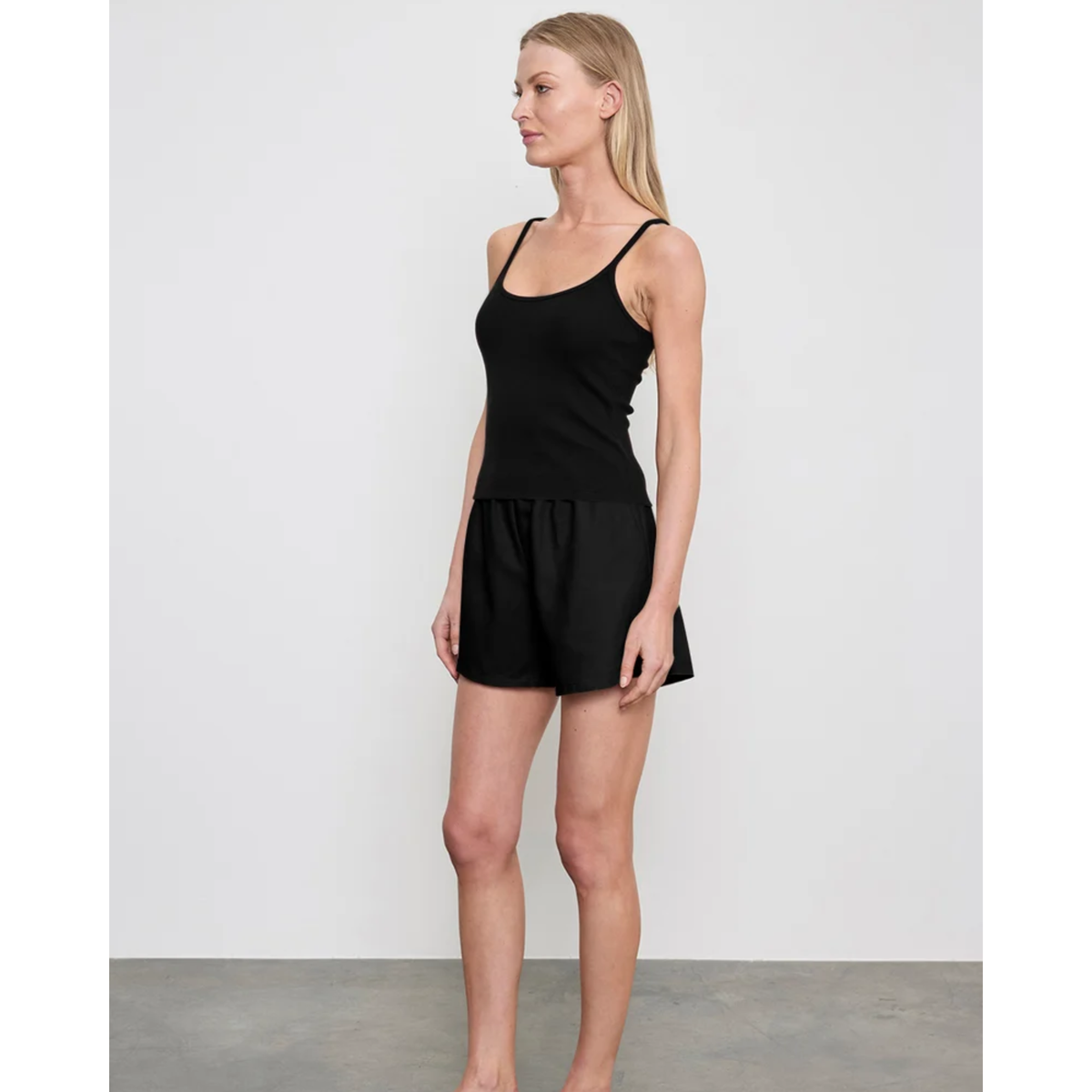 A woman modeling a black sleeveless camisole-style top with matching black shorts against a plain light gray studio background.