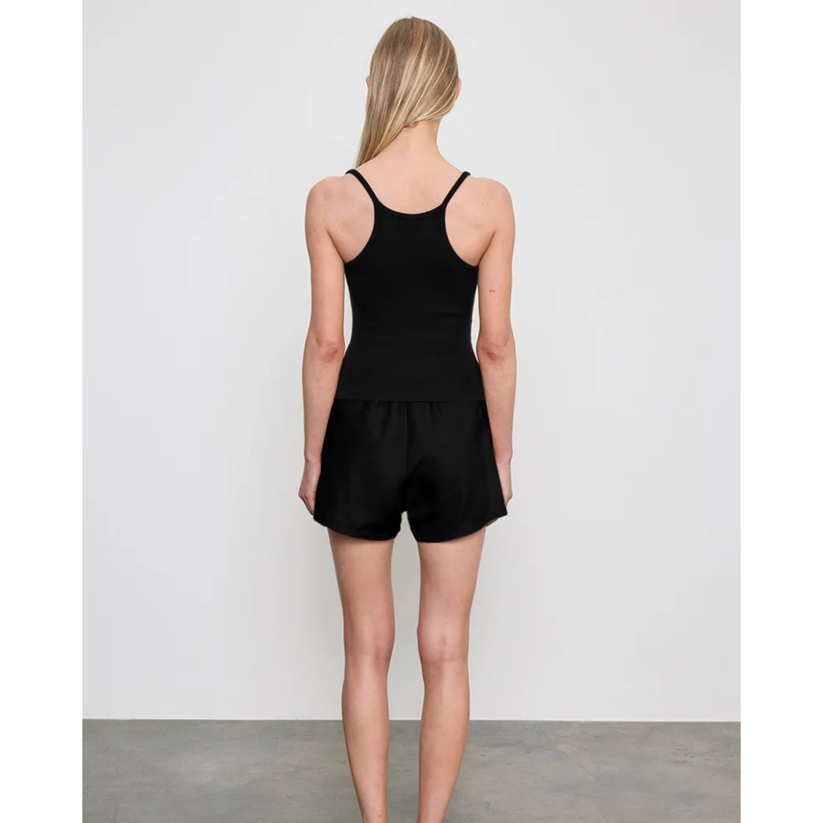 A woman viewed from the back wearing a black sleeveless romper with thin shoulder straps and short legs, standing against a plain light gray wall.