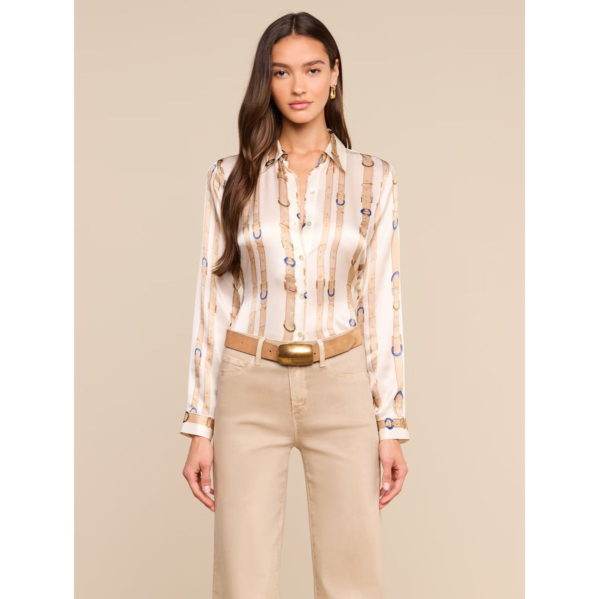 A woman modeling a long-sleeve button-up shirt with a beige and white vertical stripe pattern featuring small blue and gold graphic details, paired with beige pants and a tan belt, against a solid beige background.