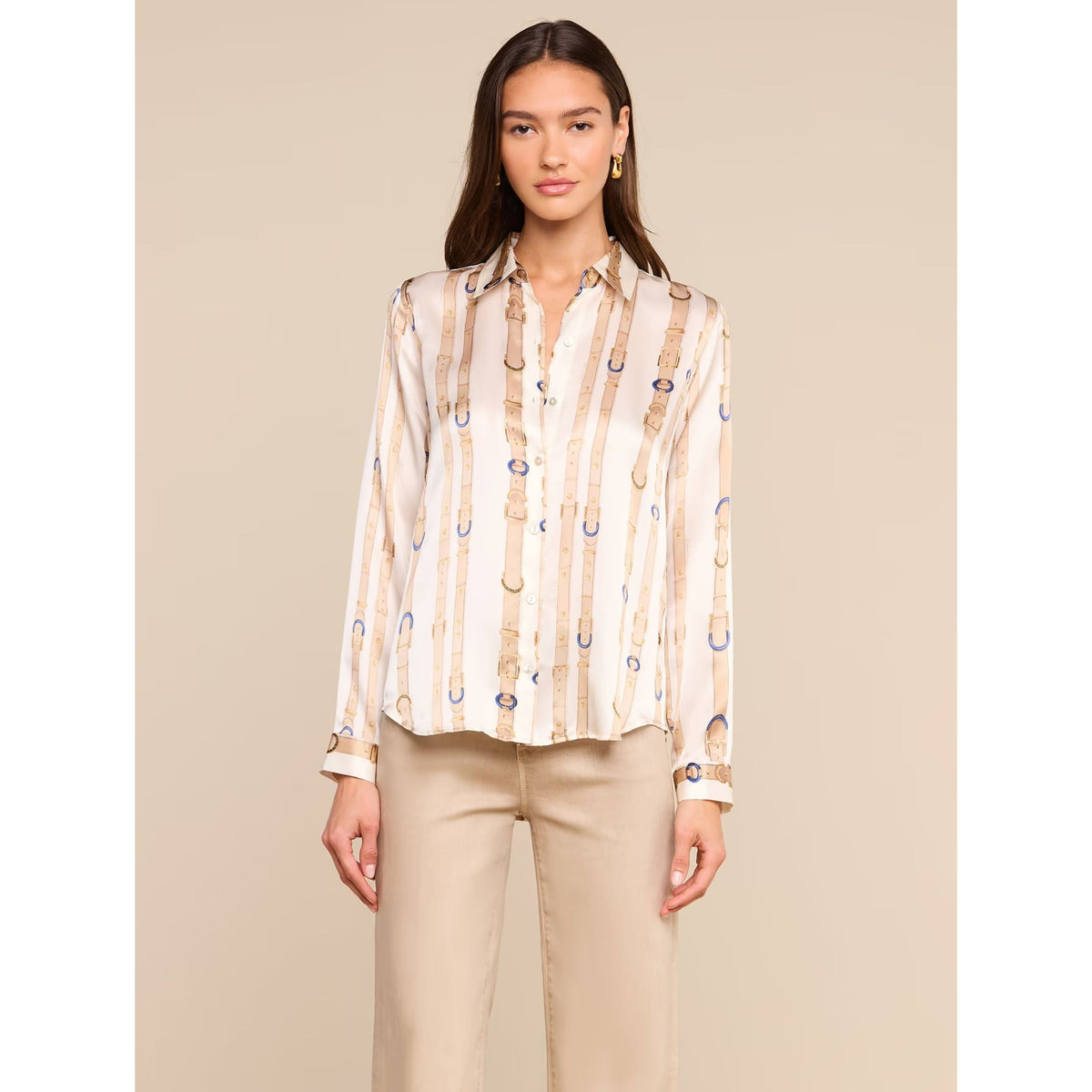 A woman modeling a long-sleeve button-up shirt with vertical beige and white stripes and blue horseshoe motifs, paired with beige pants, against a solid beige background.