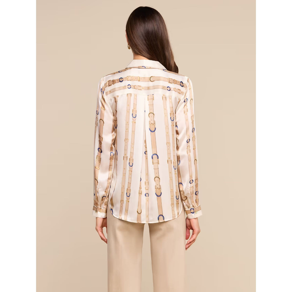 A woman viewed from the back wearing a cream satin shirt with vertical gold chain-link patterns and blue ring accents, paired with beige trousers, against a solid beige background.