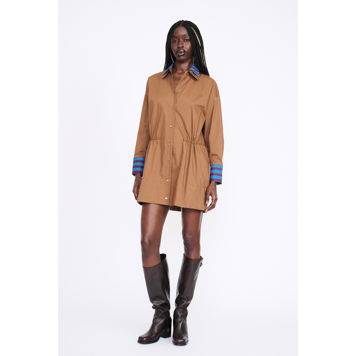A model wearing a tan button-up shirt dress with blue striped cuffs and dark brown knee-high boots, standing against a plain white studio background.