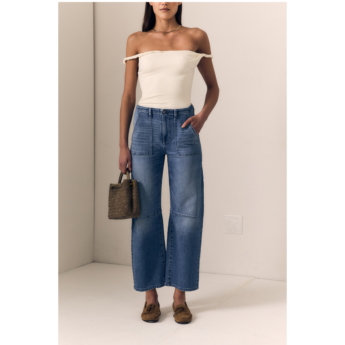 A model wearing an off-the-shoulder cream top and high-waisted blue wide-leg patchwork jeans, holding a brown woven handbag and wearing brown suede loafers, standing in a minimalist beige room.