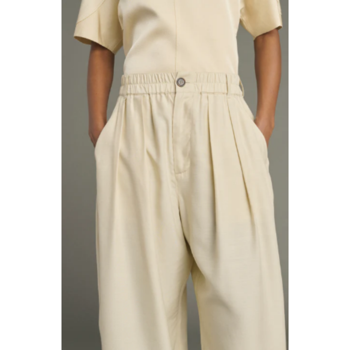 A person wearing cream-colored wide-leg trousers with a pleated front and an elastic waistband, paired with a matching cream short-sleeve top, shown against a plain gray background.