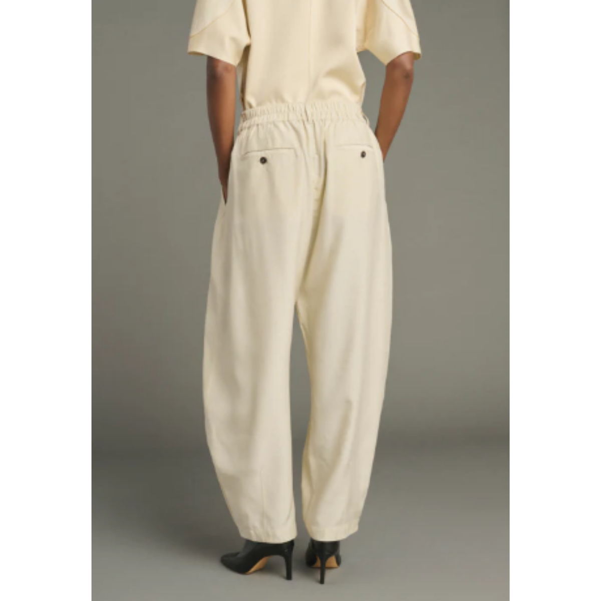 A person shown from the back wearing cream-colored wide-leg trousers with an elastic waistband and buttoned back pockets, paired with black high heels against a plain gray studio background.