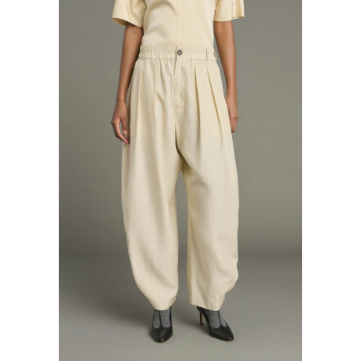 A model is wearing a pair of cream-colored, high-waisted balloon pants with a gathered elastic waistband and wide legs, paired with a matching cream top and black pointed-toe heels, set against a plain gray studio background.