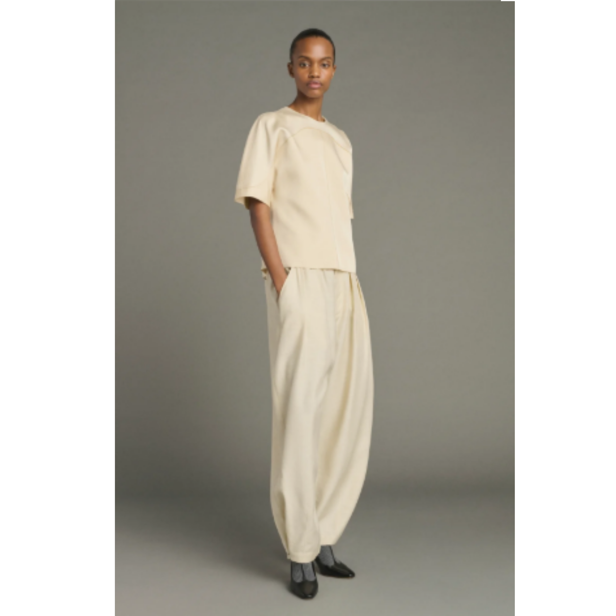 A model stands against a neutral gray studio background wearing a cream-colored two-piece outfit consisting of a short-sleeve top and matching wide-leg trousers, paired with black pointed shoes.