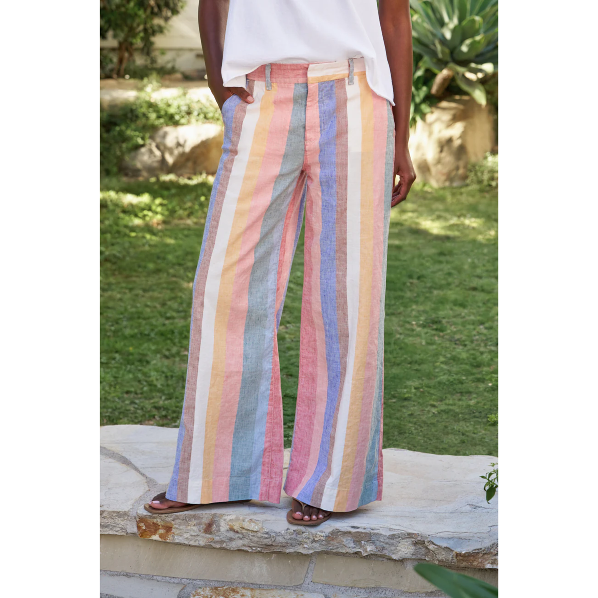 A person wearing wide-leg striped pants with vertical pastel stripes in pink, blue, yellow, and white, paired with a white top and brown sandals, standing outdoors on a stone ledge with grass in the background.