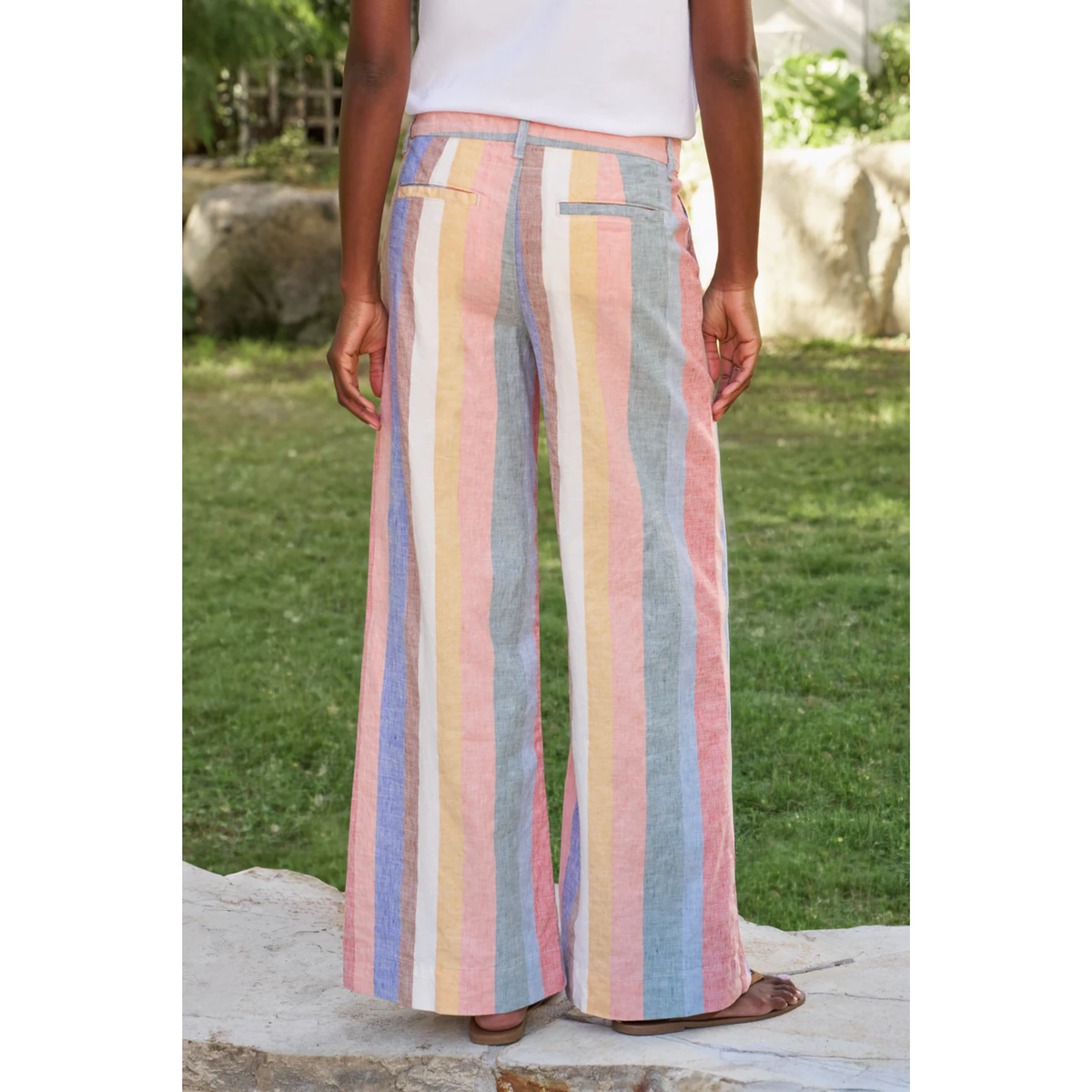A person standing outdoors on a stone surface wearing pastel vertical-striped wide-leg pants with a white top. The pants feature stripes in pink, light blue, pale yellow, and off-white.