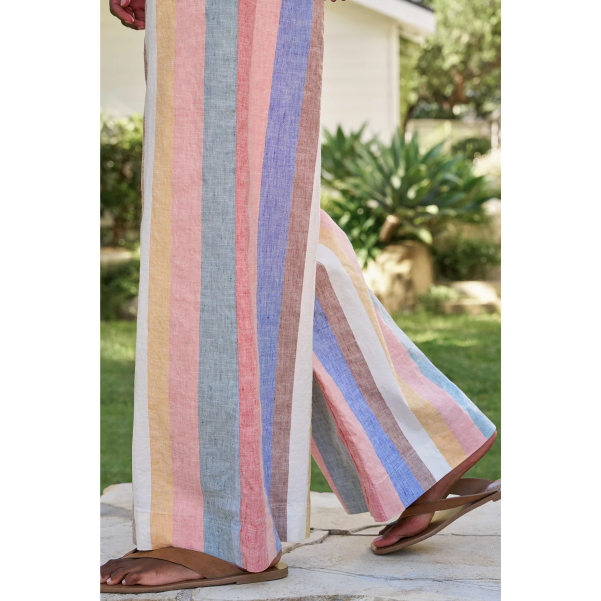 A person wearing wide-leg linen pants with vertical pastel stripes in pink, blue, yellow, and brown, paired with brown flip-flop sandals in an outdoor garden setting.