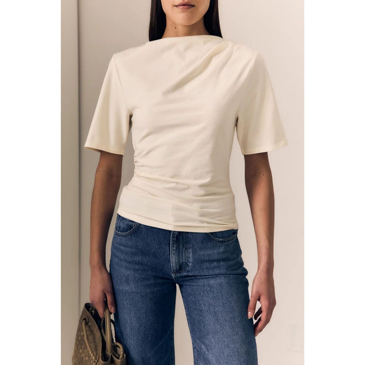 A woman wearing a cream-colored short-sleeve top with a draped neckline and gathered waist, paired with blue jeans, holding a brown woven handbag.