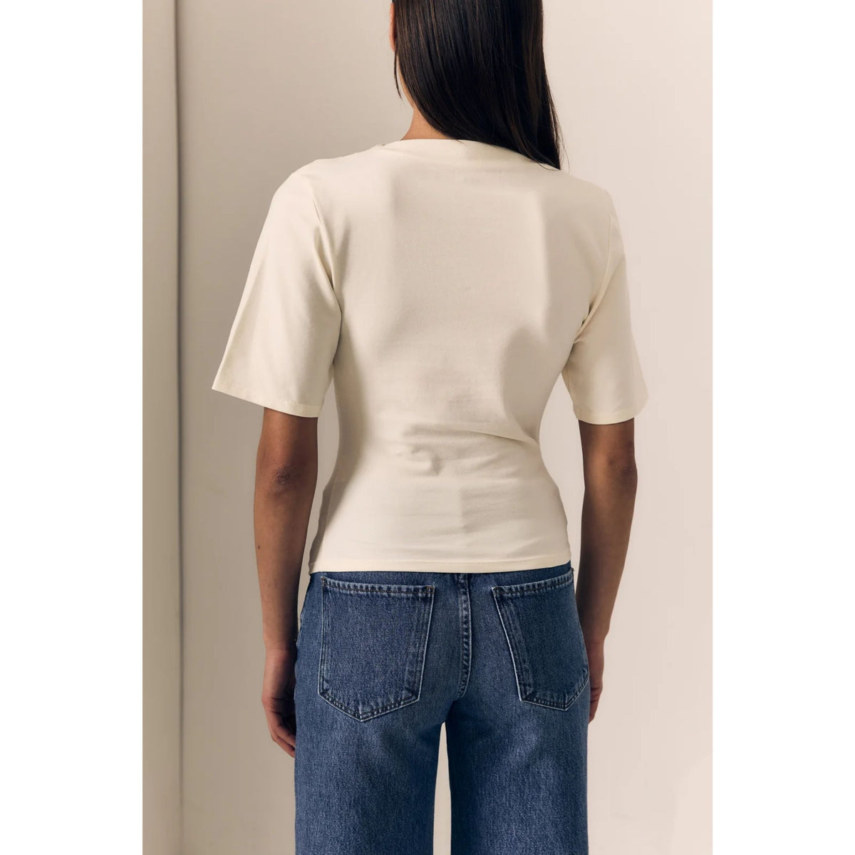 A woman viewed from the back wearing a fitted short-sleeve cream t-shirt and blue jeans, standing against a plain light beige wall.