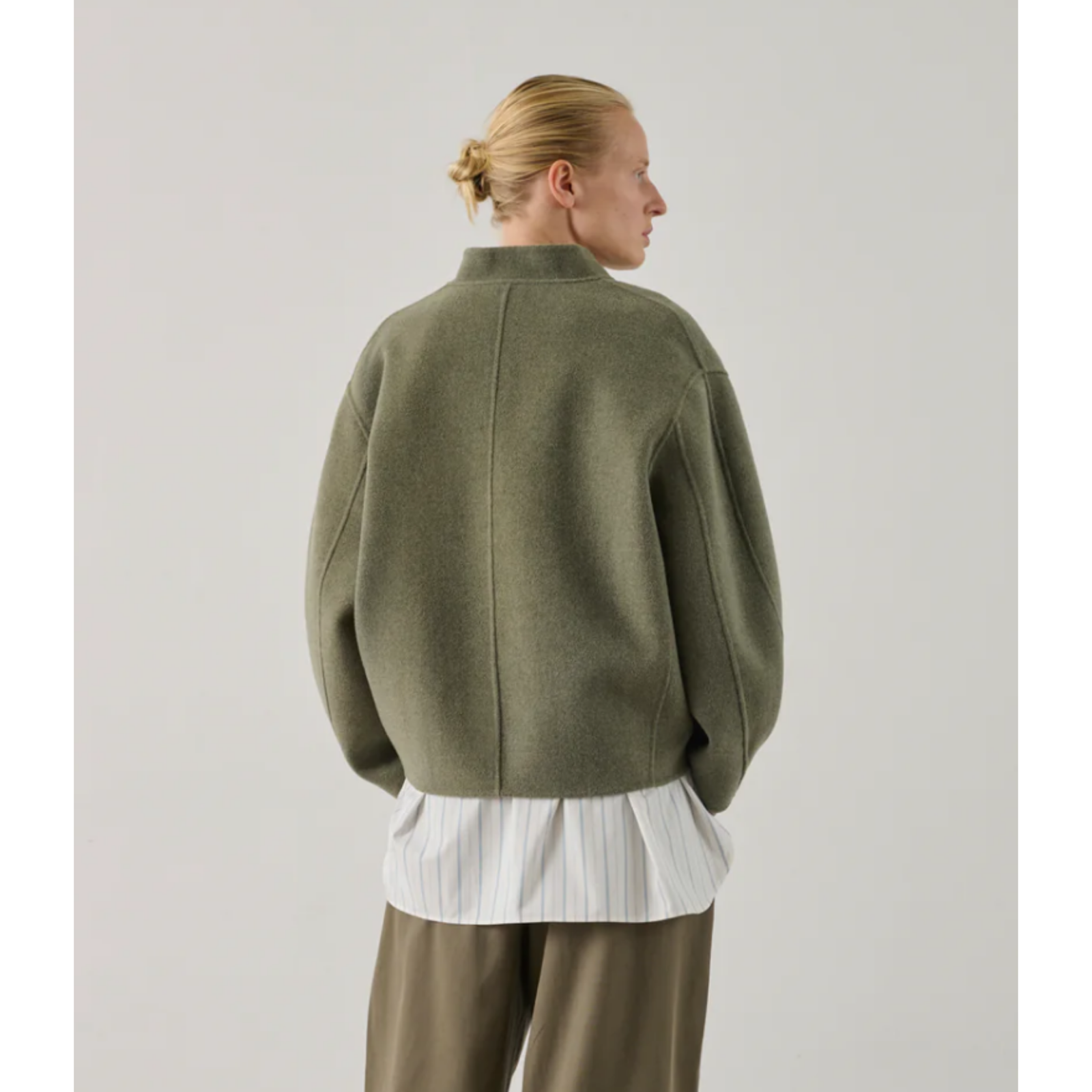 A model stands in a studio wearing an olive green cropped button-front cardigan with large front patch pockets, layered over a white striped shirt and paired with wide-leg olive pants.