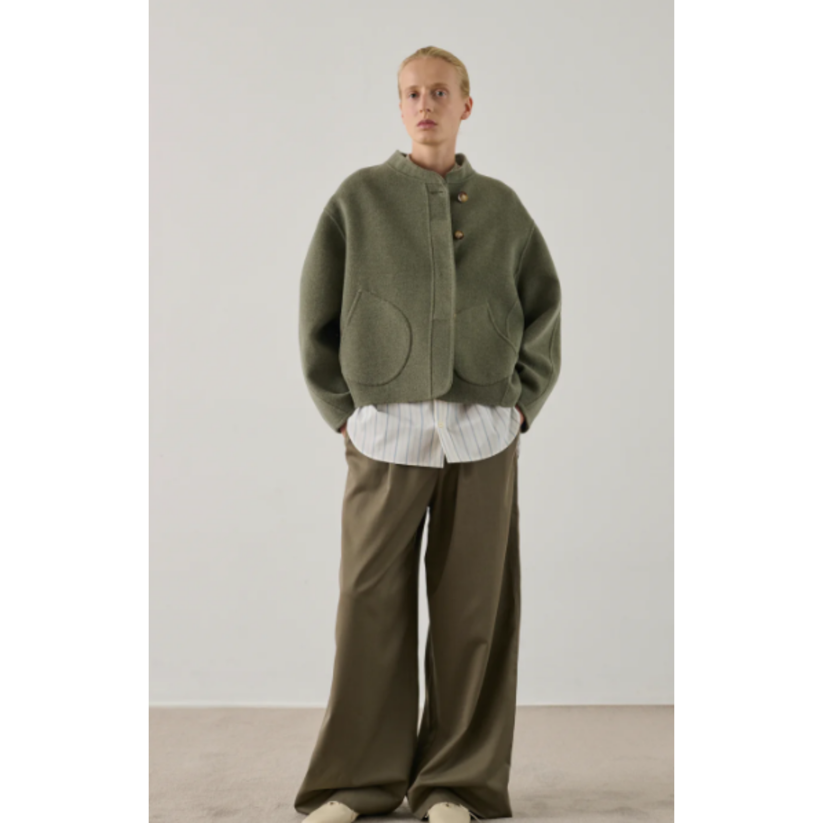 A model stands in a studio wearing an olive green cropped button-front cardigan with large front patch pockets, layered over a white striped shirt and paired with wide-leg olive pants.