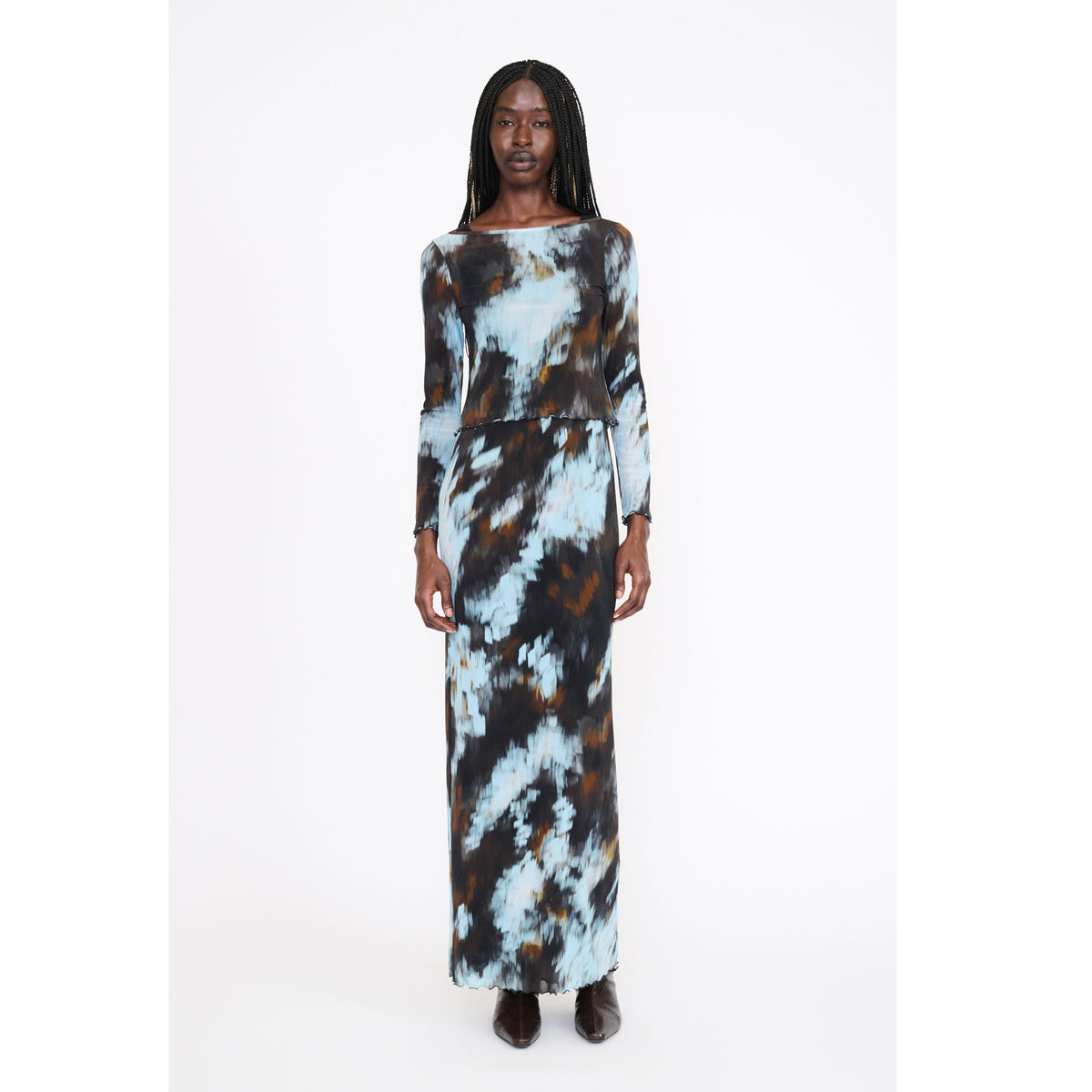 A woman modeling a long-sleeved maxi dress with a tie-dye pattern in light blue, brown, and black, standing against a plain white studio background.