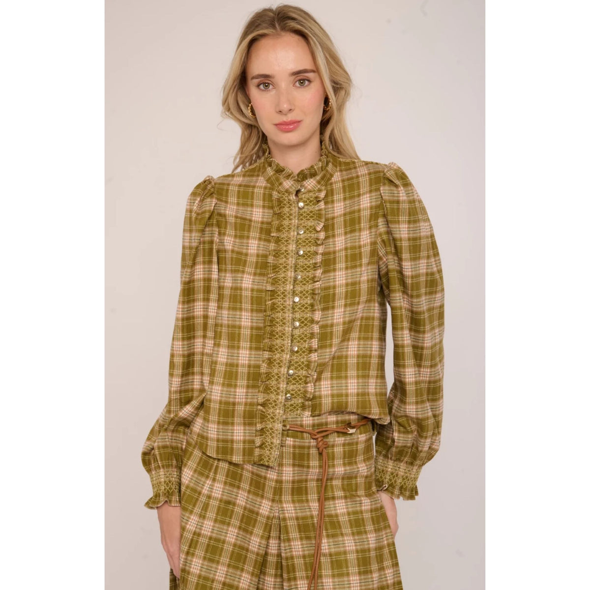 A woman modeling a long-sleeve button-front blouse with a high collar and a green and beige plaid pattern, paired with matching plaid bottoms, against a plain light background.