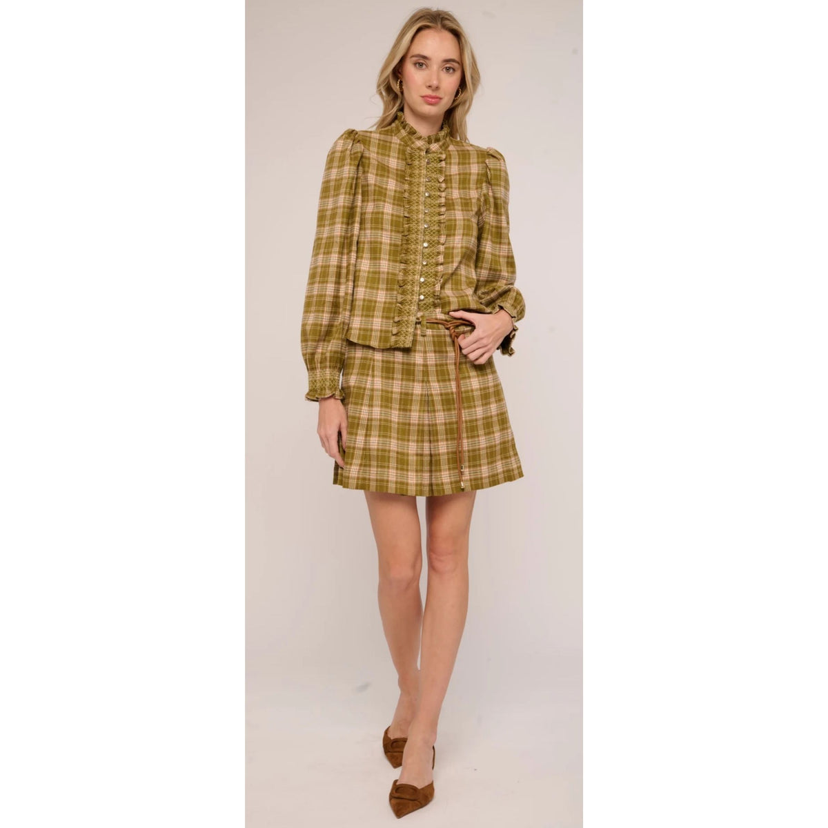 A woman modeling a matching plaid long-sleeve blouse and mini skirt set in a mustard yellow and green check pattern, paired with brown pointed-toe heels, against a plain light beige studio background.