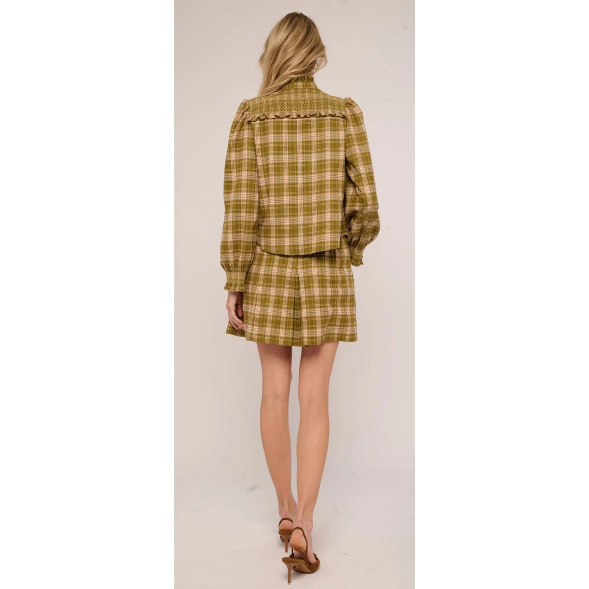 A woman is shown from the back wearing a matching plaid long-sleeve blouse and mini skirt set in olive green and beige, paired with brown high-heeled sandals, against a plain light beige studio background.