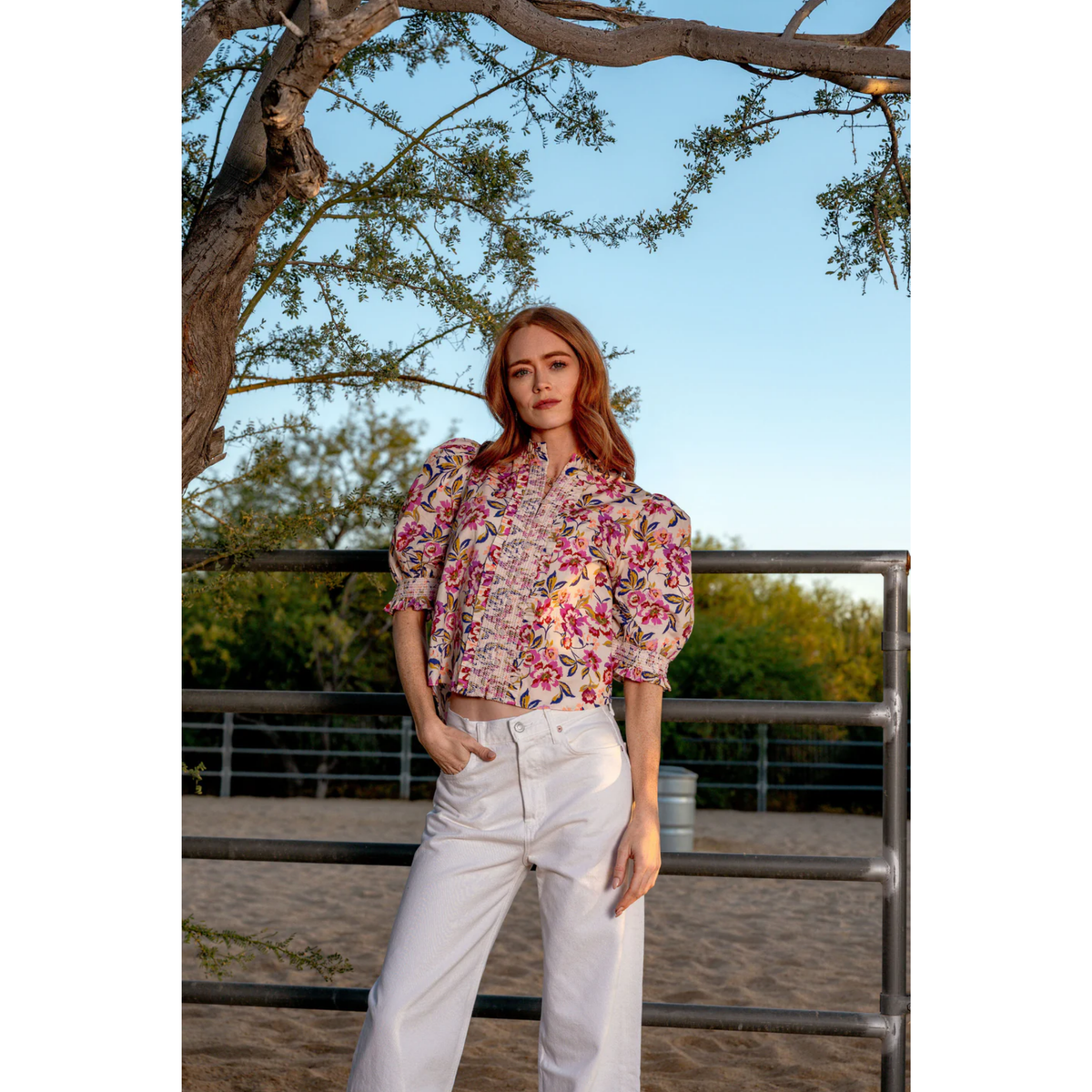 Woman wearing a floral blouse and white pants standing outdoors with trees in the background