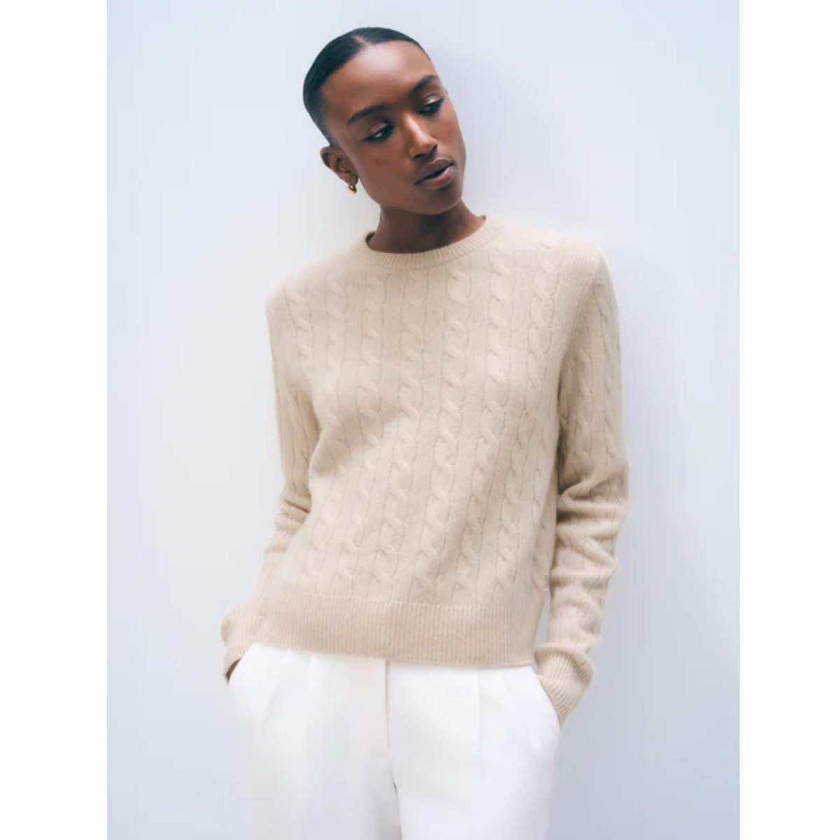 A model wearing a beige cable-knit crewneck sweater and white trousers, standing against a light gray studio background.