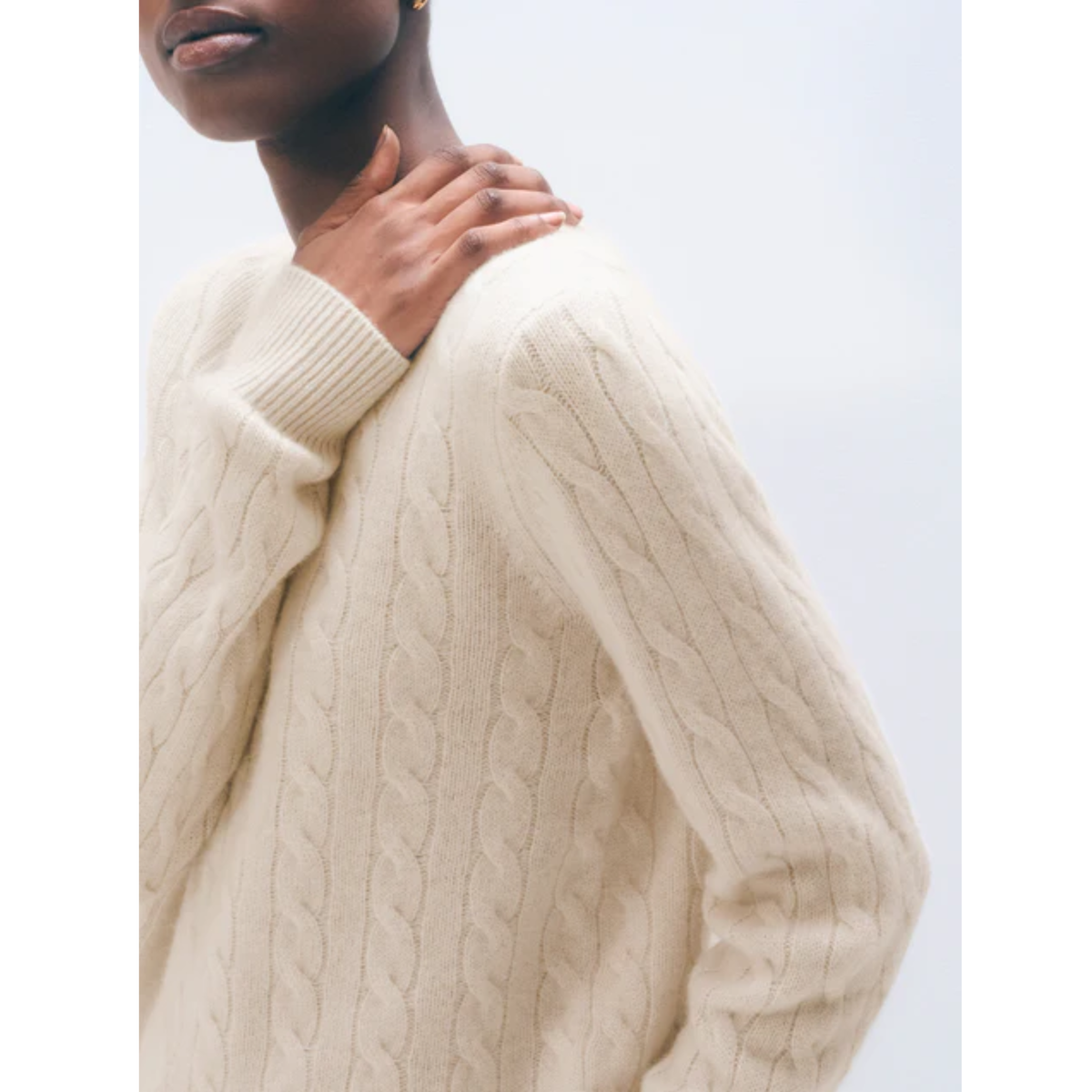 A close-up of a person wearing a cream cable-knit sweater against a light gray background.