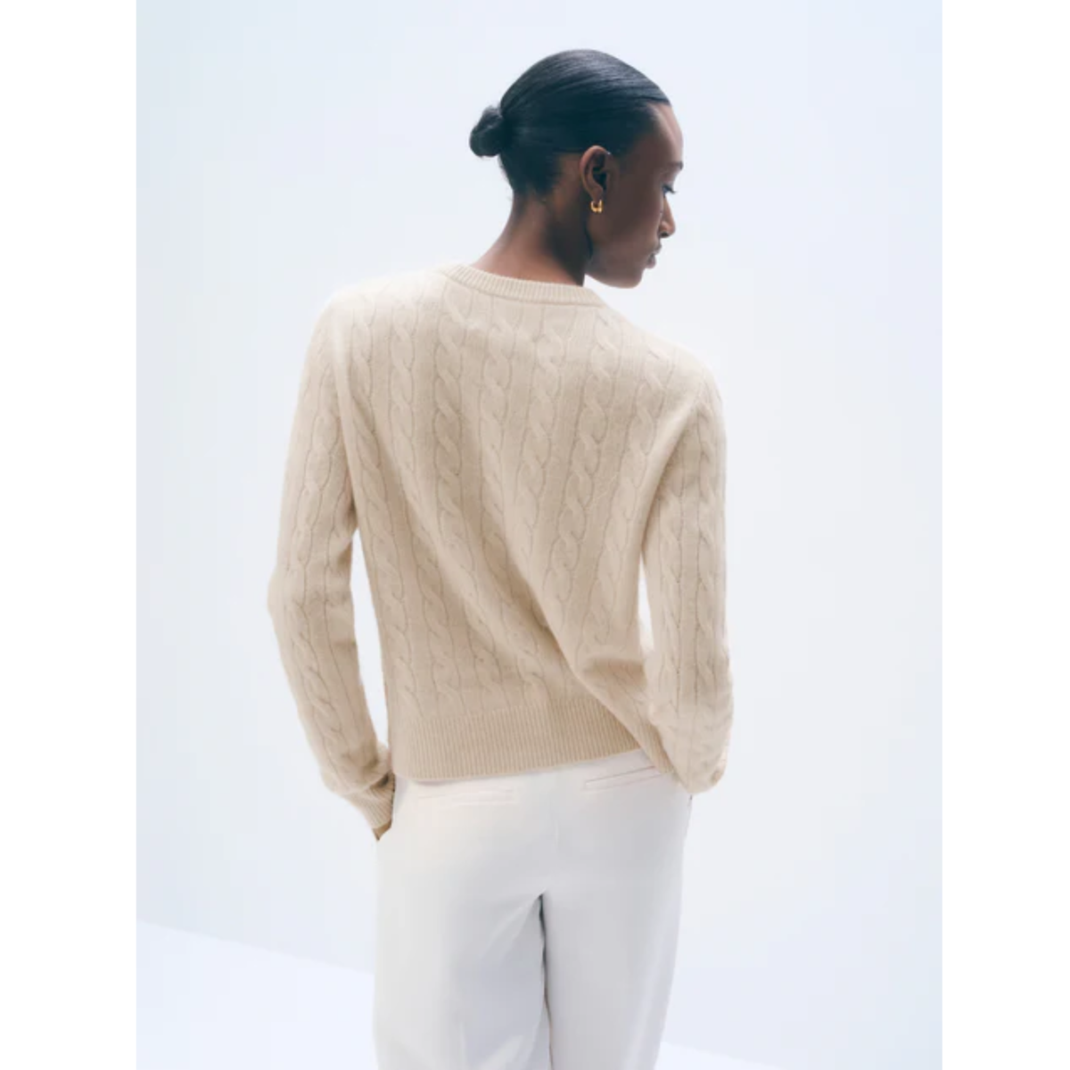 A woman shown from the back wearing a cream cable-knit sweater and white trousers against a plain light background.