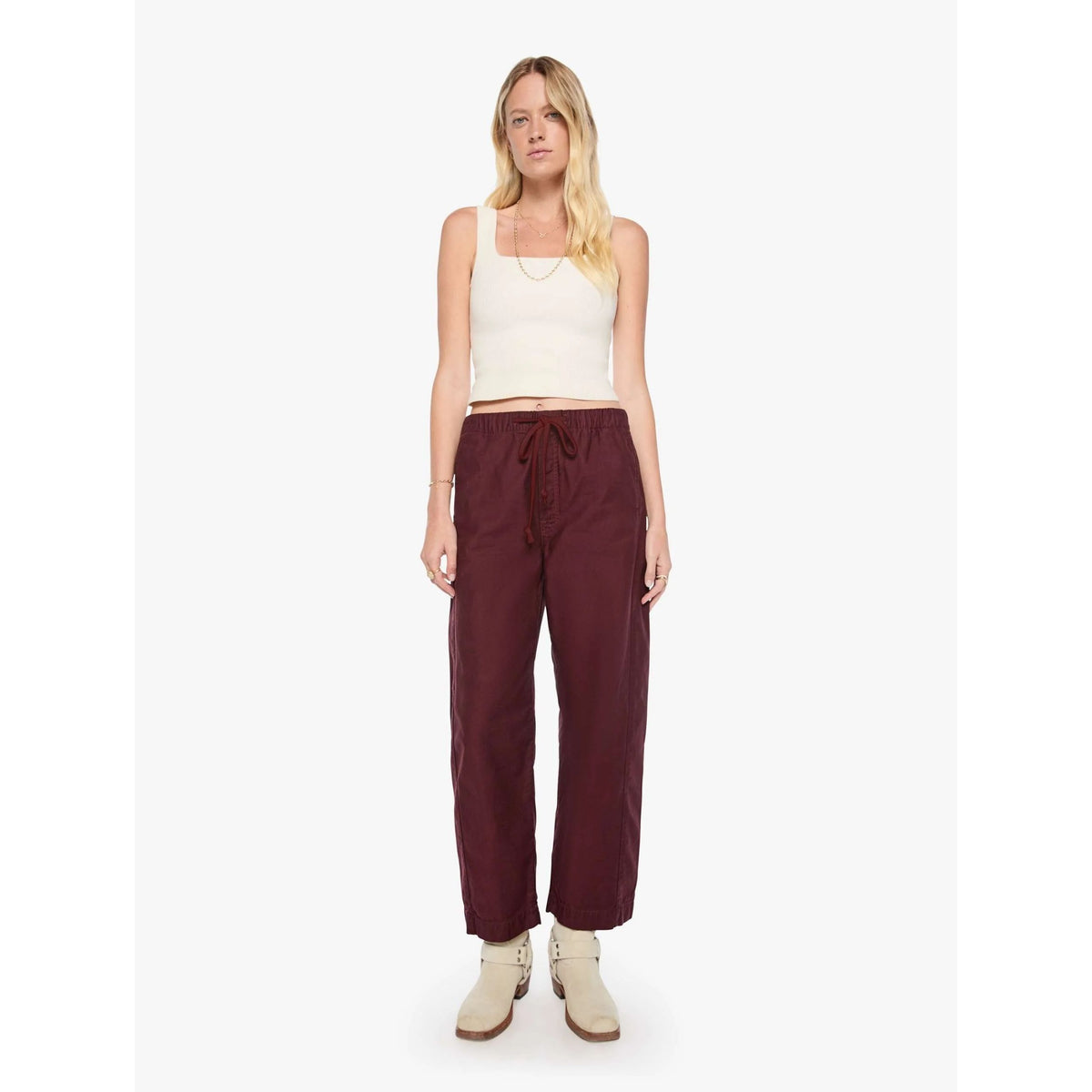 A woman modeling a pair of dark burgundy drawstring wide-leg pants, paired with a cream ribbed tank top and beige ankle boots, against a plain white studio background.