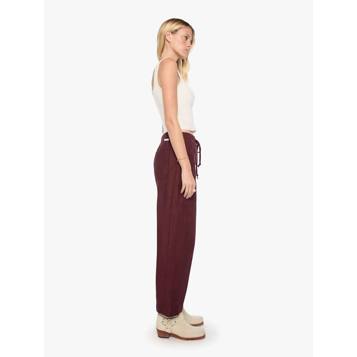 A woman shown in profile wearing loose, wide-leg burgundy drawstring pants, a white ribbed tank top, and cream ankle boots against a plain light gray studio background.