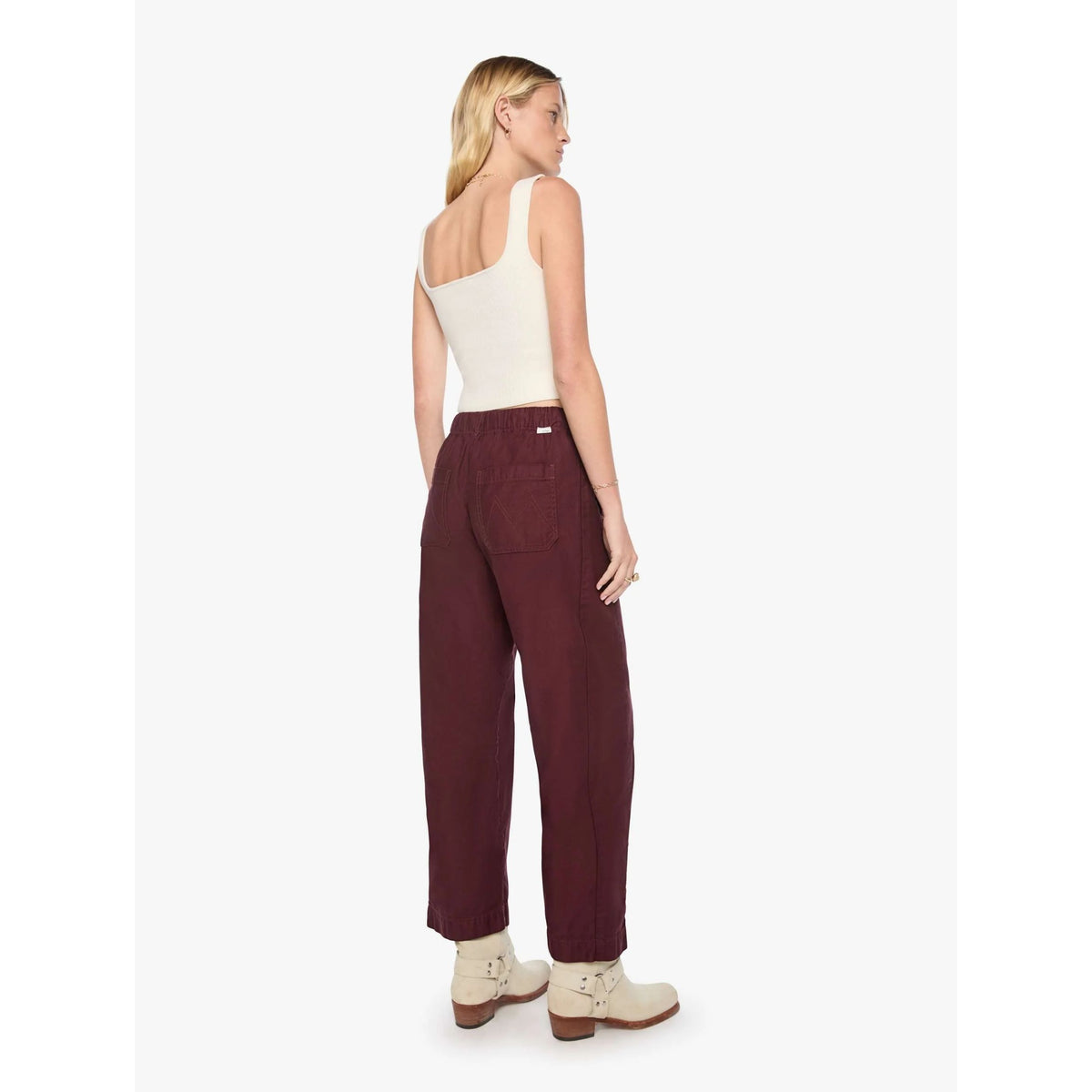 A woman shown from the back wearing burgundy wide-leg pants, a cream tank top, and cream ankle boots against a plain light gray studio background.