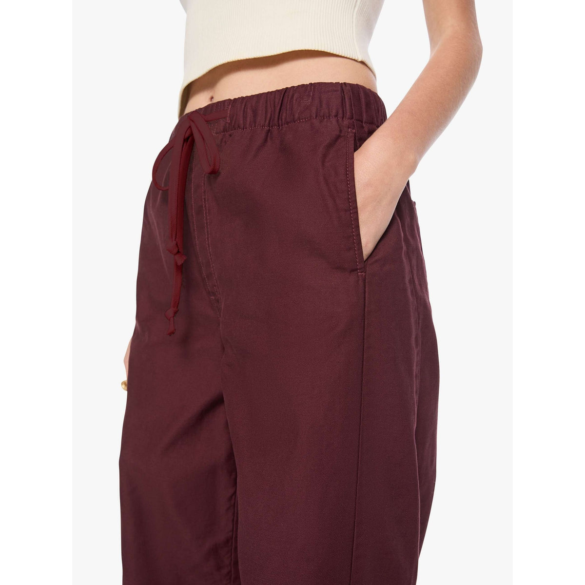 A close-up of a person wearing loose, wide-leg burgundy drawstring pants with side pockets, paired with a cream ribbed crop top against a plain light background.