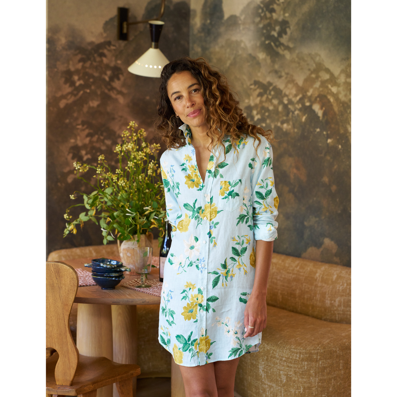 Woman wearing a floral dress standing in a room with a table and plants.
