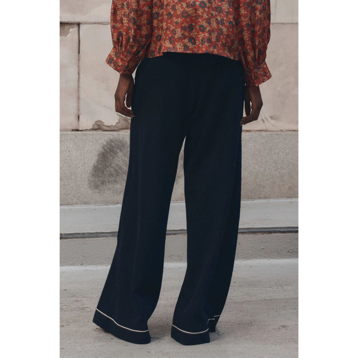 A person shown from the back wearing dark navy wide-leg trousers with thin white piping along the hem, paired with a rust-orange floral blouse, standing outdoors against a light stone wall.