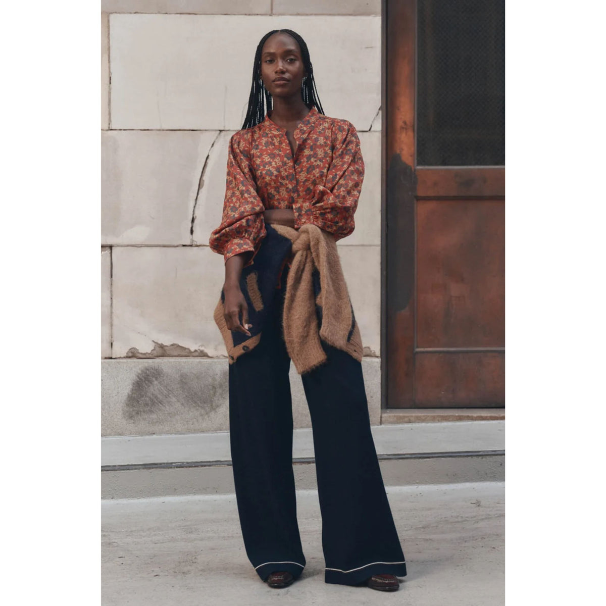 A woman stands outdoors in front of a stone wall and wooden door, wearing a cropped orange floral blouse with long sleeves, dark wide-leg pants, and a tan jacket draped over one arm.