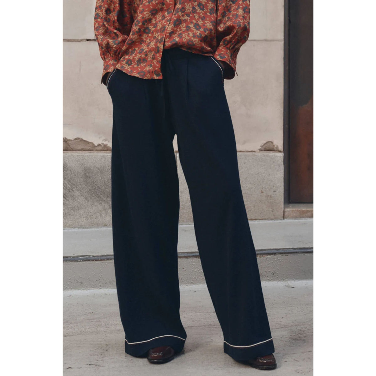 A person wearing dark navy wide-leg trousers with thin white piping along the hem, paired with a rust-orange floral blouse and brown shoes, standing outdoors against a concrete wall.