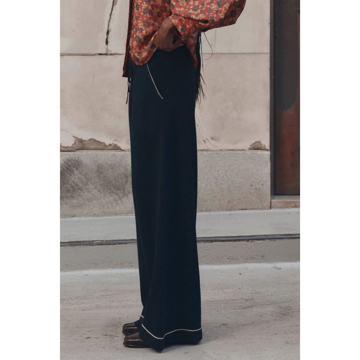 A person wearing dark navy wide-leg trousers with thin light piping along the hem, paired with a rust-orange floral blouse and brown loafers, standing outdoors against a concrete wall.