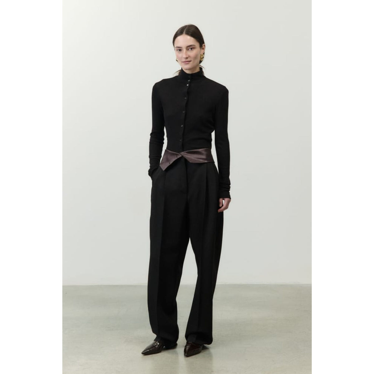 A model stands in a studio wearing a black long-sleeve button-front top with a high mock neck, black wide-leg trousers, and dark brown pointed shoes. A dark brown satin-like sash is tied at the waist.